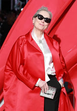 A woman wearing a bright red satin coat over a white satin blouse and black pants, with black sunglasses and a small metallic clutch, standing against a large red backdrop.