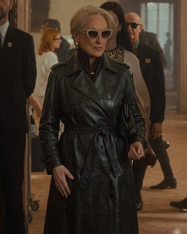 A woman wearing a black leather trench coat and white cat-eye sunglasses walks through a hallway with other people in the background.