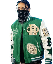 A person wearing a green and white varsity-style jacket with large embroidered letter patches, ribbed green and white sleeves, and a black bandana covering the face, photographed in a blue-lit event setting.