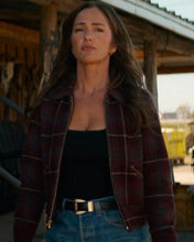 A woman wearing a dark red and black plaid cropped jacket over a black top and blue jeans, standing in front of a rustic wooden structure.