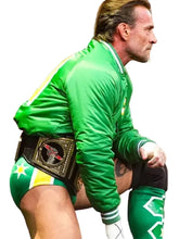 A wrestler in a bright green tracksuit and matching green-and-white shorts, wearing a large championship belt around his waist, captured mid-action on a blurred stadium background.
