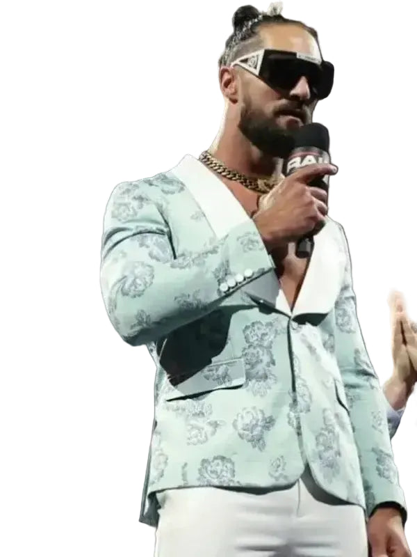 A man in a light mint green patterned blazer and white pants holding a microphone on stage, wearing black sunglasses and a gold chain necklace.