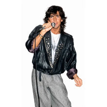 A man wearing a black studded leather jacket over a white t-shirt and gray pants, holding a microphone against a plain white background.
