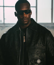 A man wearing black sunglasses and a black leather bomber jacket with a Stone Island patch on the pocket, standing in front of a bright window.