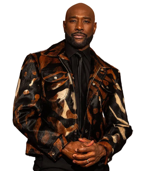 A man wearing a black, brown, and beige patterned jacket over a black shirt and tie, standing against a solid warm brown background.