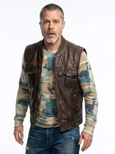 A man wearing a dark brown leather motorcycle vest over a camouflage long-sleeve shirt and blue jeans, standing against a plain white background.
