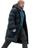 A man with red hair and a beard wearing a long black puffer coat over a teal shirt and dark trousers, captured in motion against a plain white background.