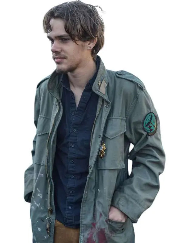 A man wearing a distressed olive green military-style jacket over a dark navy button-down shirt, with a gold pin on the chest pocket, set against a plain white background.
