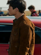 A man in profile wearing a brown suede jacket, standing outdoors next to a red car with a blurred background.