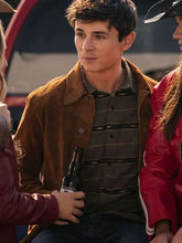 A man wearing a brown suede jacket over a dark striped polo shirt, holding a beer bottle in a casual outdoor setting with other people partially visible.