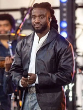 A man on a stage wearing a black leather jacket over a white collared shirt and light blue jeans, holding a microphone.