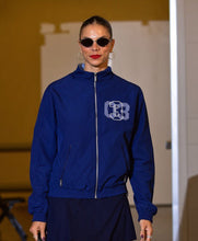 A woman wearing a royal blue zip-up jacket with a white and blue 'B' monogram on the chest, styled with dark sunglasses and hoop earrings, standing against a warm beige background.