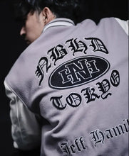 A close-up back view of a person wearing a light gray varsity-style jacket with large black embroidered text and an oval 'HNI' logo on the back, against a dark background.