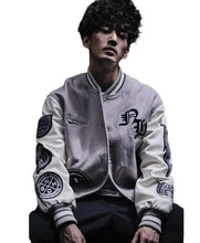 A man wearing a light gray and white varsity jacket with black patches and letter embroidery, posed against a dark background.