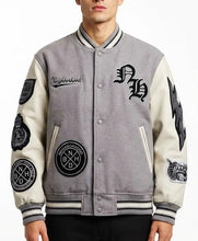 A man wearing a gray and cream varsity jacket with multiple black embroidered patches and lettering, including 'Neighborhood' and 'NMD'.