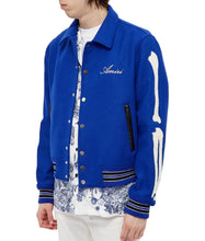 Blue varsity jacket with 'Amiri' branding worn by a person on a white background
