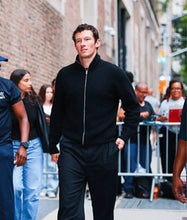 A man walking outdoors in a black zip-up jacket and black pants, photographed on a city street with buildings and other people visible in the background.