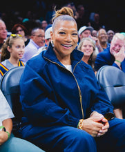 A smiling woman seated in an arena crowd, wearing a dark blue zip-front jacket and matching bottoms with gold zipper accents.