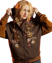 A woman wearing a brown varsity-style jacket with tan sleeves and hood, decorated with colorful embroidered autumn motifs including pumpkins, leaves, and a fox, posed against a dark background.