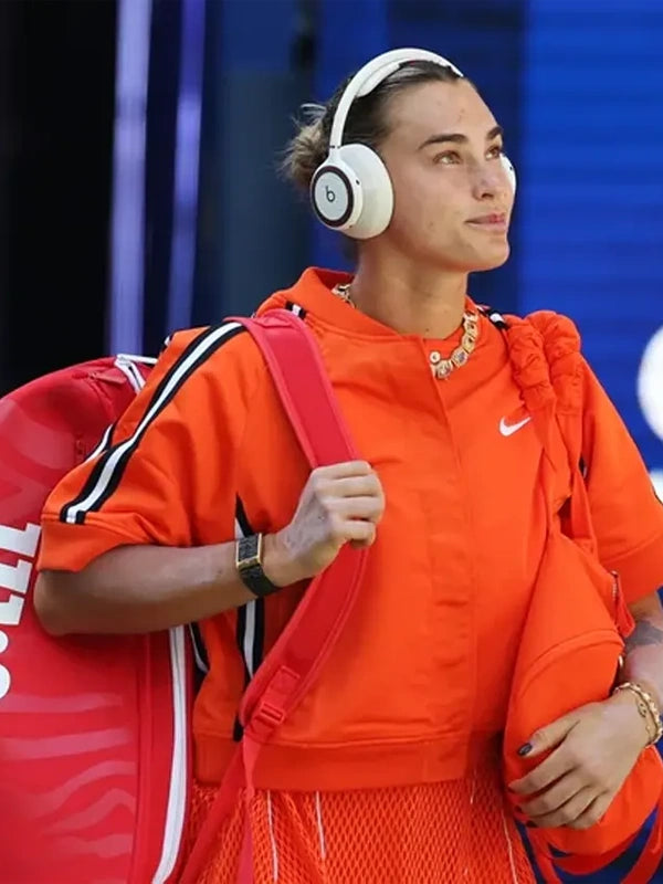 A woman in a bright orange athletic tracksuit wearing white Beats headphones and carrying a red tennis bag, captured in a real event setting against a blue background.