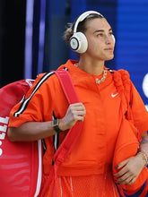 A woman in a bright orange athletic tracksuit wearing white Beats headphones and carrying a red tennis bag, captured in a real event setting against a blue background.