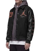 A person wearing a black varsity-style jacket with leather-look sleeves, a gray hoodie underneath, and brown Jordan number and logo graphics on the chest and sleeve.