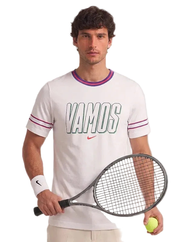 A man modeling a white Nike t-shirt with 'VAMOS' printed across the chest in green and white, accented with purple and pink stripes on the collar and sleeves. He holds a tennis racket and a tennis ball against a solid gray background.
