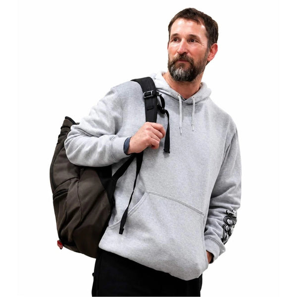 A man wearing a light gray hoodie and black pants, carrying a dark brown backpack over his shoulder, against a solid white background.