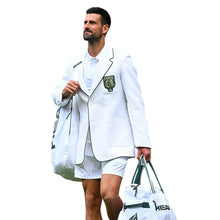 A man in a white blazer and white shorts walks on a tennis court carrying white tennis bags. The blazer has green trim and a crest patch on the left chest.