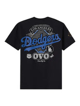 A black short-sleeve t-shirt with a large Los Angeles Dodgers graphic on the back, featuring blue and white lettering and logos, displayed against a light gray background.