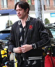 A man wearing a black satin bomber jacket with white stripes and a red devil patch on the chest, standing outdoors on a city street.