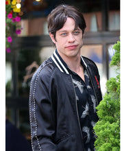 A man wearing a black bomber jacket with white stripes on the collar and sleeves, layered over a black shirt with a white bird pattern, standing outdoors in front of a blurred building background.