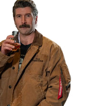 A man with a mustache wearing a brown Old Forester work jacket and holding a glass of amber liquid, set against a dark, industrial background.