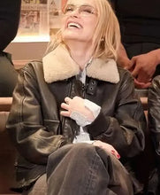 A woman wearing a black leather aviator-style jacket with a cream shearling collar, sitting in an indoor setting and laughing while wearing clear-framed sunglasses.