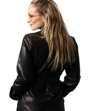 A woman with long blonde hair in a high ponytail, wearing glasses and a black leather jacket, shown from a side-back angle against a plain white background.