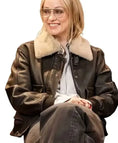 A woman wearing a dark brown leather jacket with a thick beige shearling-style collar, a light gray button-up shirt, and dark gray pants, photographed against a plain white background.