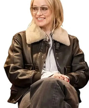 A woman wearing a dark brown leather jacket with a thick beige shearling-style collar, a light gray button-up shirt, and dark gray pants, photographed against a plain white background.