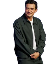 A man wearing a dark olive green jacket over a white t-shirt, standing in front of a blue event backdrop with white text.