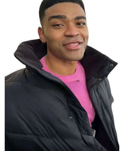 A man wearing a black puffer jacket over a bright pink t-shirt, shown from the chest up against a plain white background.