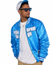 A man wearing a shiny light blue varsity-style bomber jacket with 'GREETYS' and 'TICKS TO' embroidered on the front, paired with a white t-shirt, khaki pants, and a white cap. A woman in a matching outfit is partially visible on the left against a blue-lit event background.