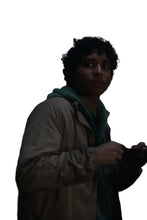 A man in a beige jacket and green hoodie looking off-camera in a dimly lit setting, with a large blurred sign in the background.