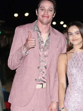 A man in a light pink blazer over a patterned pink and green shirt gives a thumbs-up, standing next to a woman in a sparkling light pink dress at a night event.
