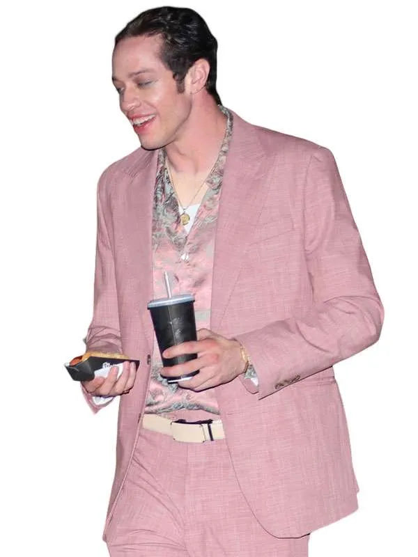 A man wearing a light pink textured blazer and matching trousers, styled with a floral-patterned shirt and a beige belt, holding a black travel tumbler and a phone against a plain white background.