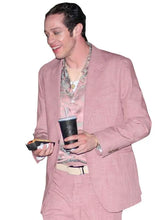 A man wearing a light pink textured blazer and matching trousers, styled with a floral-patterned shirt and a beige belt, holding a black travel tumbler and a phone against a plain white background.