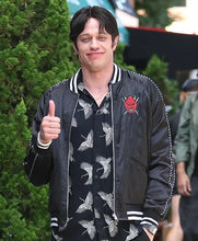 A man wearing a black satin bomber jacket with white stripes, a red devil patch, and a black-and-white bird-patterned shirt, giving a thumbs-up outdoors.