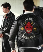 A black satin bomber jacket with a large red devil face and the word 'OKINAWA' on the back, worn by a person shown from behind. Another person in a matching jacket stands to the left.
