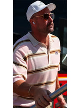 A man wearing a white baseball cap, gold-framed aviator sunglasses, and a light pink polo shirt with brown and gold horizontal stripes, holding a metal railing.