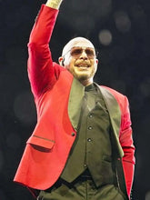 A man on stage wearing a bright red blazer over a dark olive vest and matching dark trousers, with sunglasses and one arm raised in a wave.