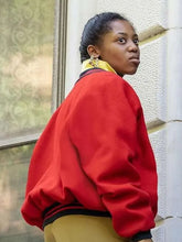A woman wearing a bright red oversized bomber jacket with black and yellow ribbed trim, paired with olive green pants, standing against a textured stone wall.