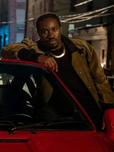 A man wearing a brown jacket and black shirt leans against a red car in a nighttime urban setting.